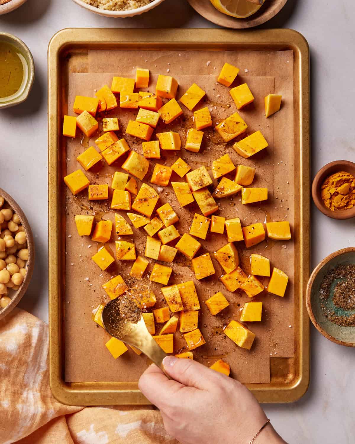 A hand sprinkles spices over cubed butternut squash on a parchment-lined baking sheet, surrounded by bowls of ingredients like chickpeas, oil, and seasonings.