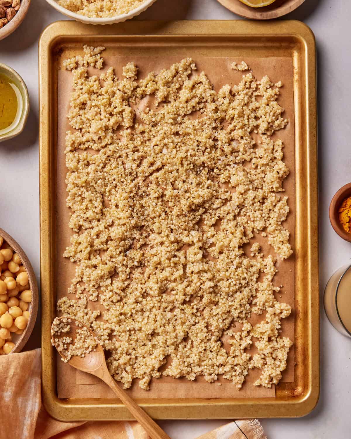 A baking sheet lined with parchment paper holds cooked quinoa spread evenly. Surrounding the tray are bowls with chickpeas, olive oil, spices, and a wooden spoon rests on the sheet.