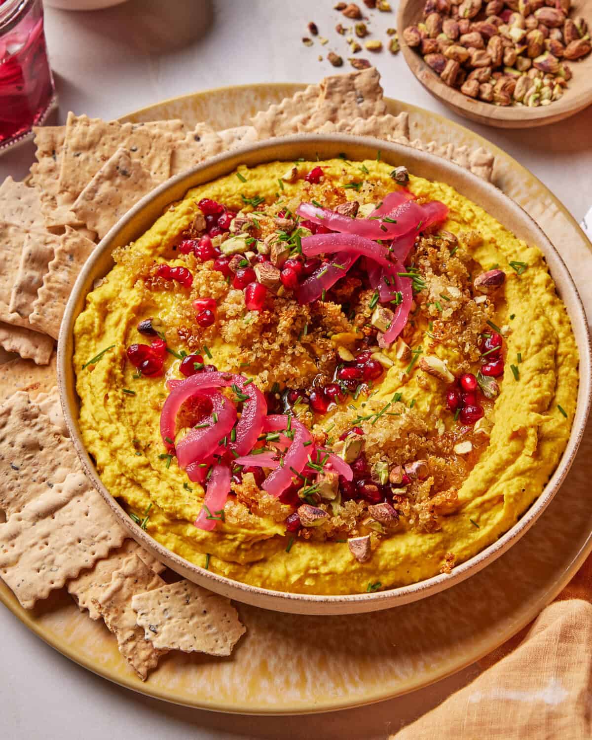 A bowl of vibrant yellow hummus topped with pickled onions, chopped pistachios, pomegranate seeds, and herbs, surrounded by flatbread crackers on a plate. A bowl of pistachios is visible in the background.