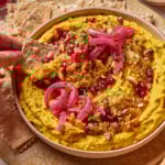 A hand dips a seeded cracker into a bowl of hummus topped with pickled onions, pomegranate seeds, chopped pistachios, herbs, and spices. Crackers are arranged around the bowl on a plate.