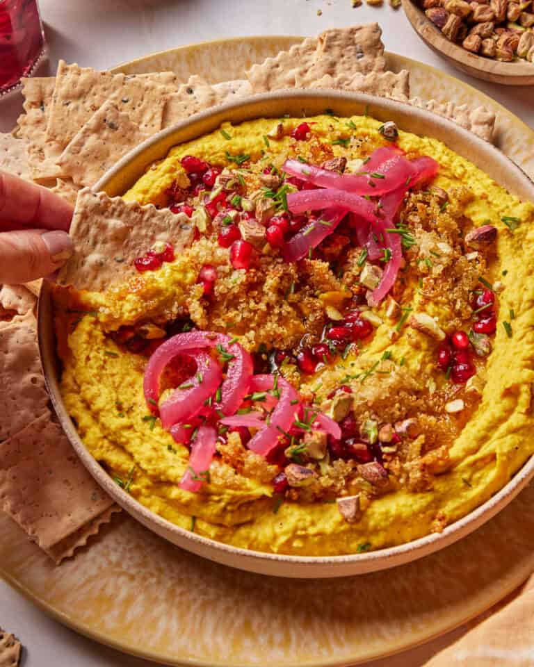 A hand dips a seeded cracker into a bowl of hummus topped with pickled onions, pomegranate seeds, chopped pistachios, herbs, and spices. Crackers are arranged around the bowl on a plate.
