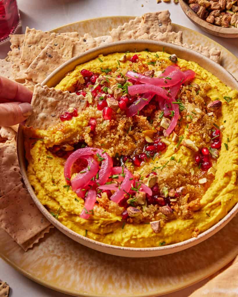 A hand dips a seeded cracker into a bowl of hummus topped with pickled onions, pomegranate seeds, chopped pistachios, herbs, and spices. Crackers are arranged around the bowl on a plate.