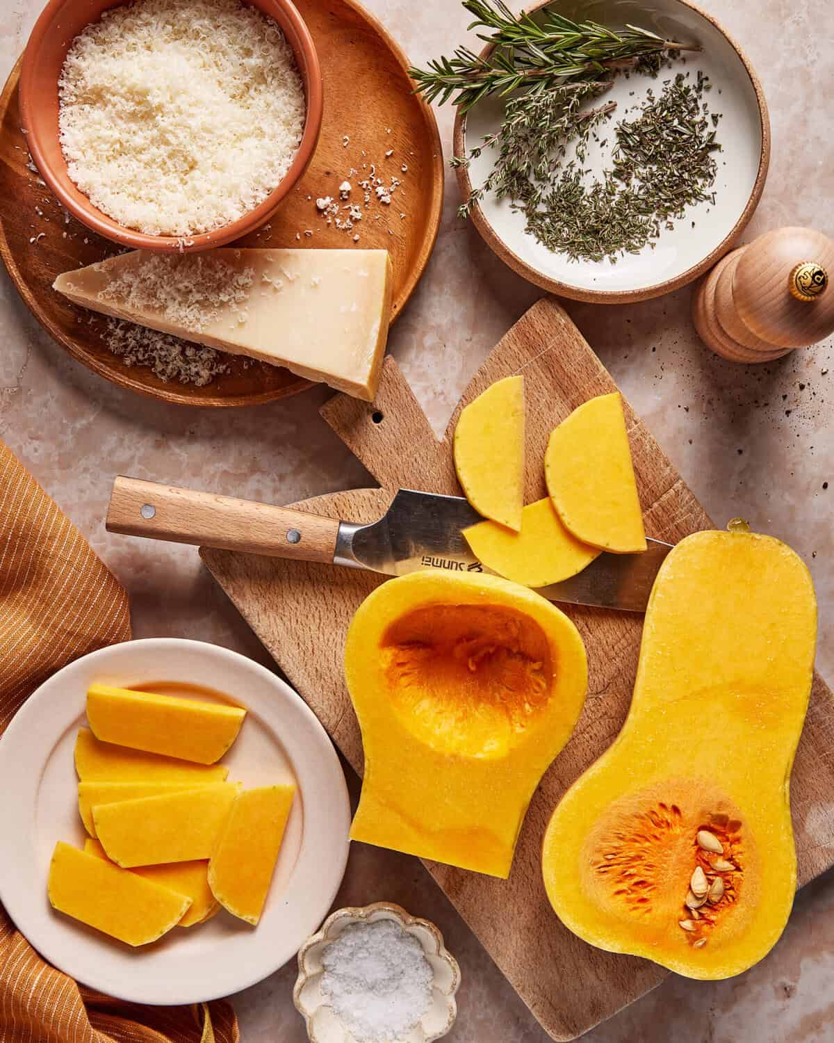 Wooden boards display sliced butternut squash, a knife, grated cheese, a wedge of parmesan, fresh rosemary, dried herbs, a pepper mill, salt, and an orange-striped cloth on a marble surface.