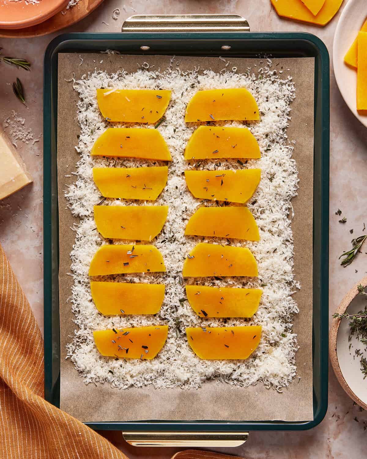 A baking tray lined with parchment paper holds rows of butternut squash slices on a bed of grated cheese, garnished with herbs. A striped cloth, fresh herbs, cheese, and squash are arranged nearby.