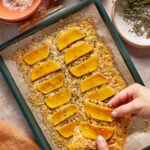 A person lifts a slice of baked cheesy flatbread topped with roasted butternut squash from a parchment-lined baking sheet; herbs, salt, and cheese are nearby.