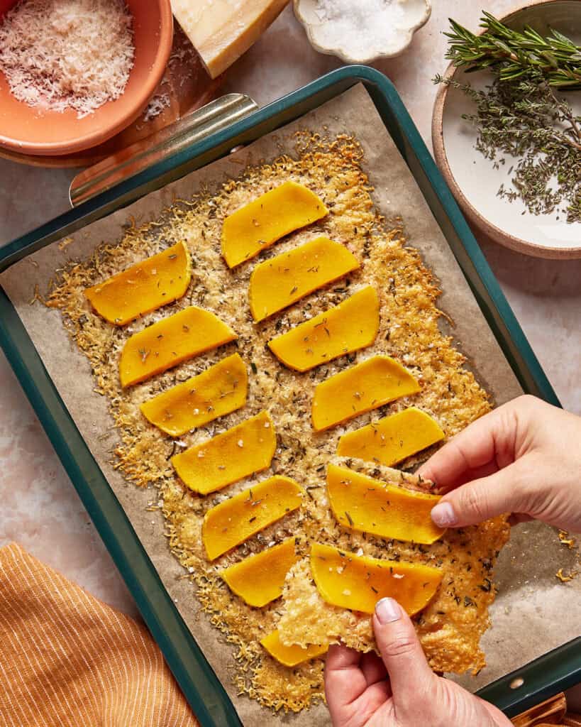 A person lifts a slice of baked cheesy flatbread topped with roasted butternut squash from a parchment-lined baking sheet; herbs, salt, and cheese are nearby.