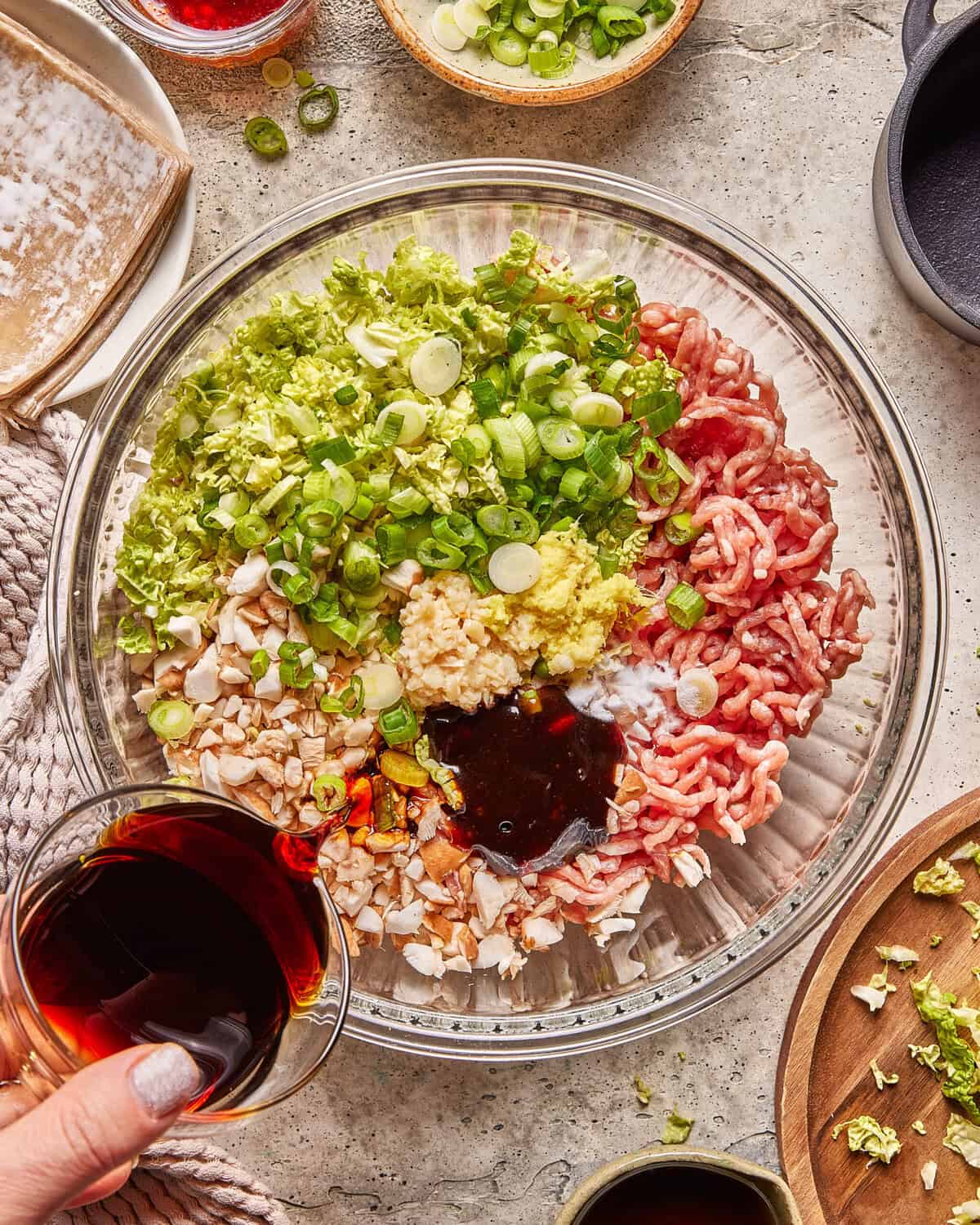 A glass bowl with ground meat, chopped lettuce, scallions, ginger, garlic, cashews, and sauce being poured in. Various ingredients and utensils surround the bowl on a light tabletop.