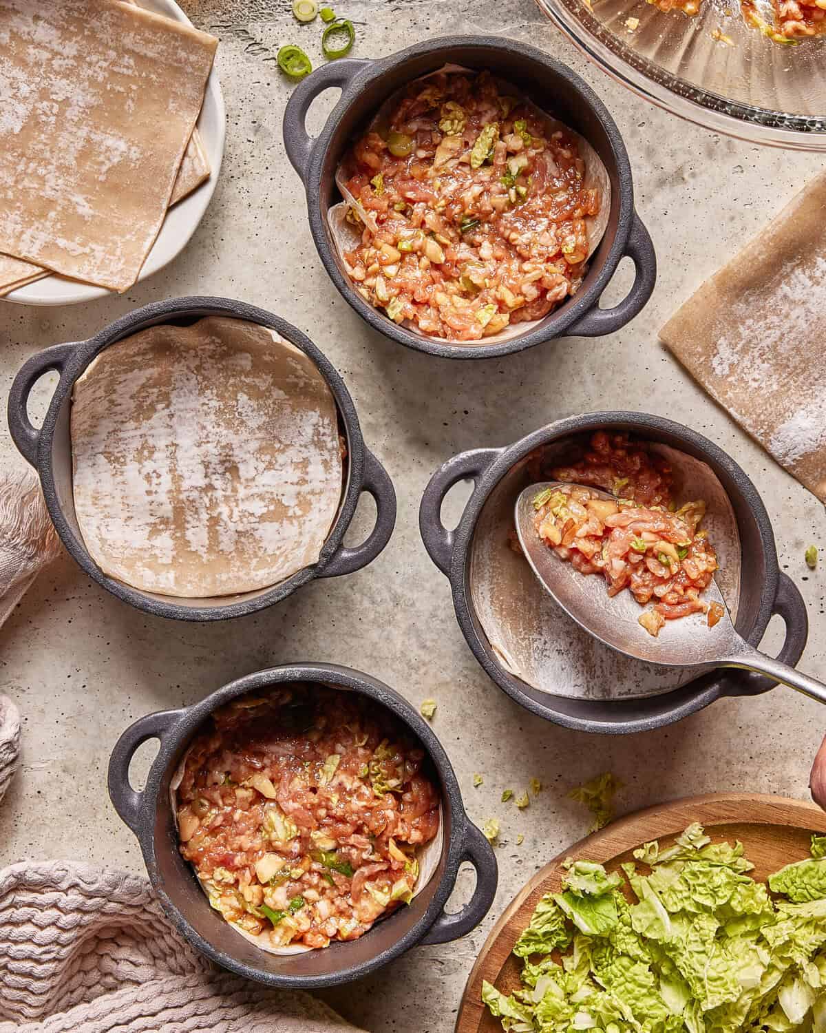 Four small cast iron pots, some lined with parchment paper, are being filled with a mixture of chopped vegetables and sauce. Unused parchment, dumpling wrappers, and cabbage pieces are nearby on a light countertop.
