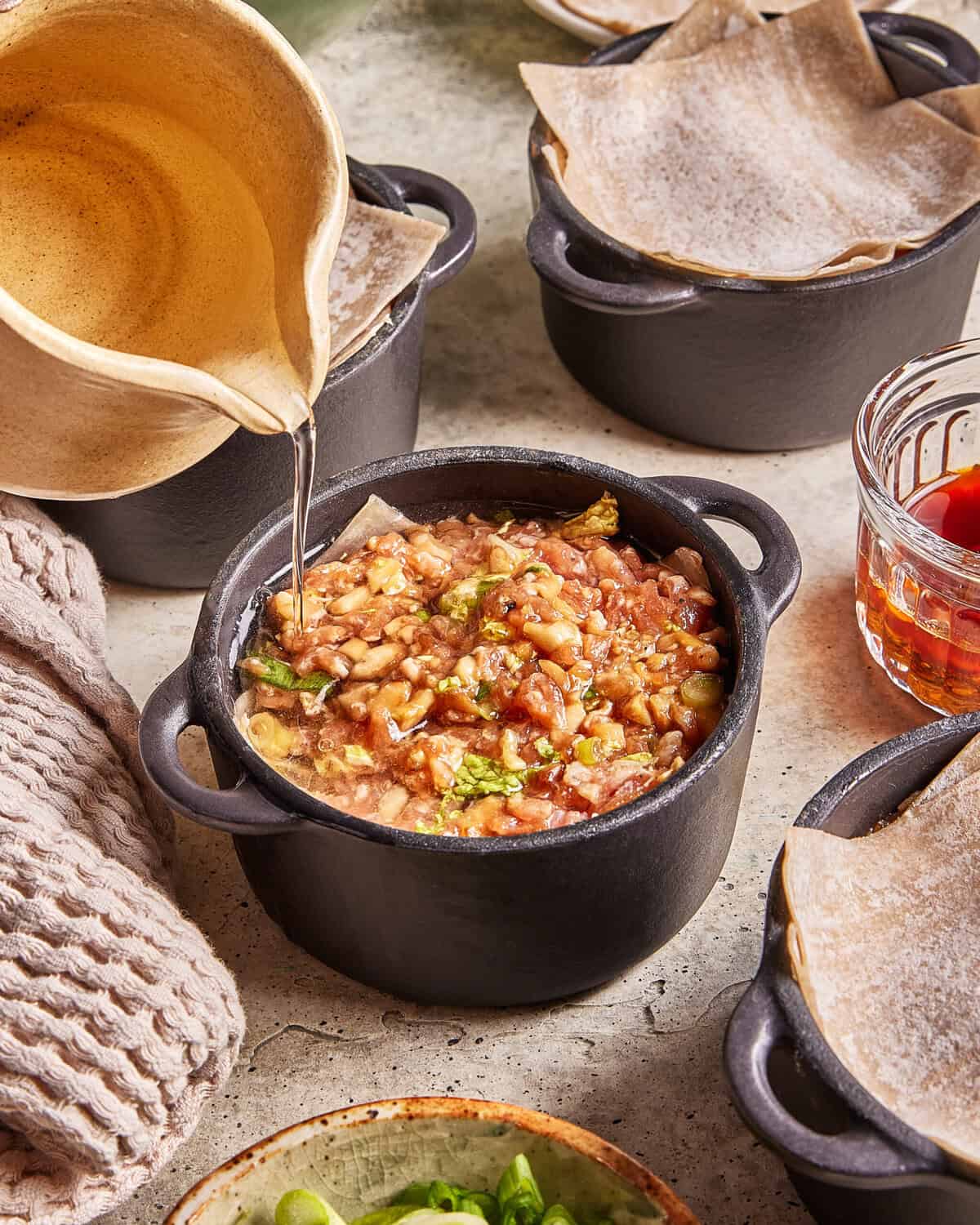 A hand pours liquid from a beige pitcher into a small black pot filled with a chunky stew, surrounded by other pots, a towel, a glass of sauce, and vegetables on a countertop.
