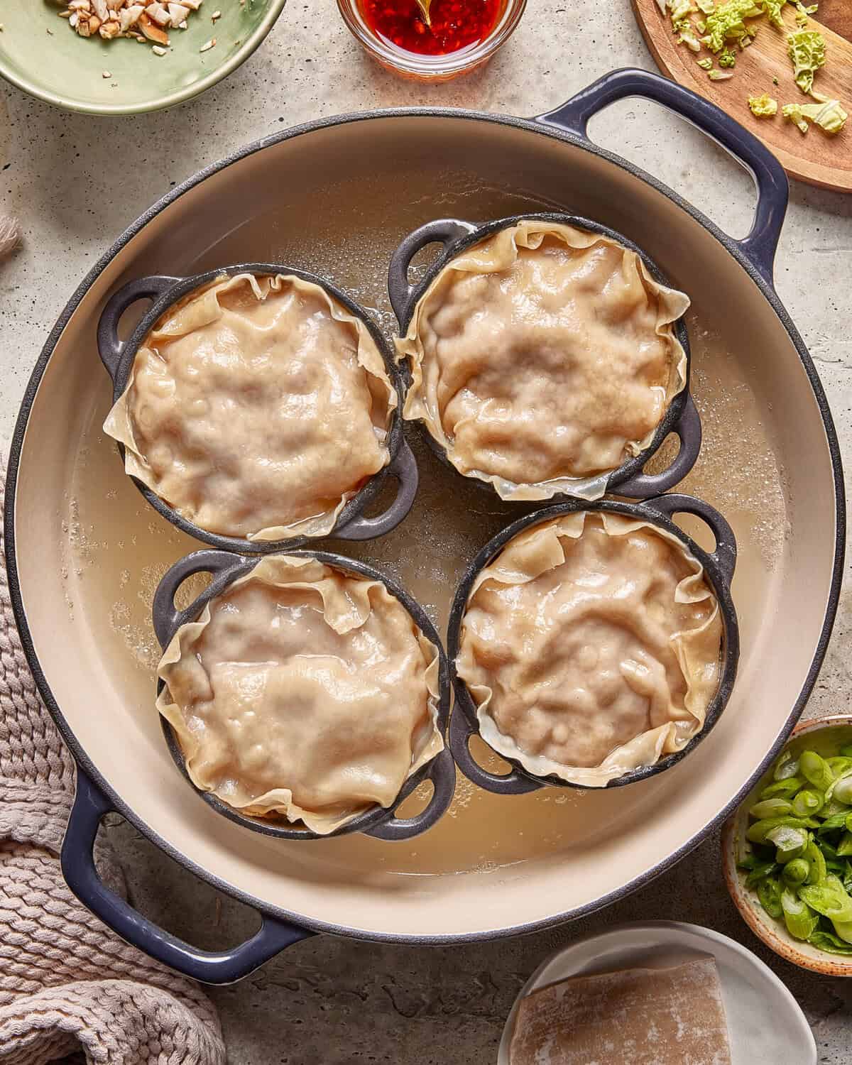 Four small round dishes filled with golden-brown pot pies are arranged in a large cream-colored pan. Various bowls with chopped vegetables and sauce surround the pan on a textured surface.