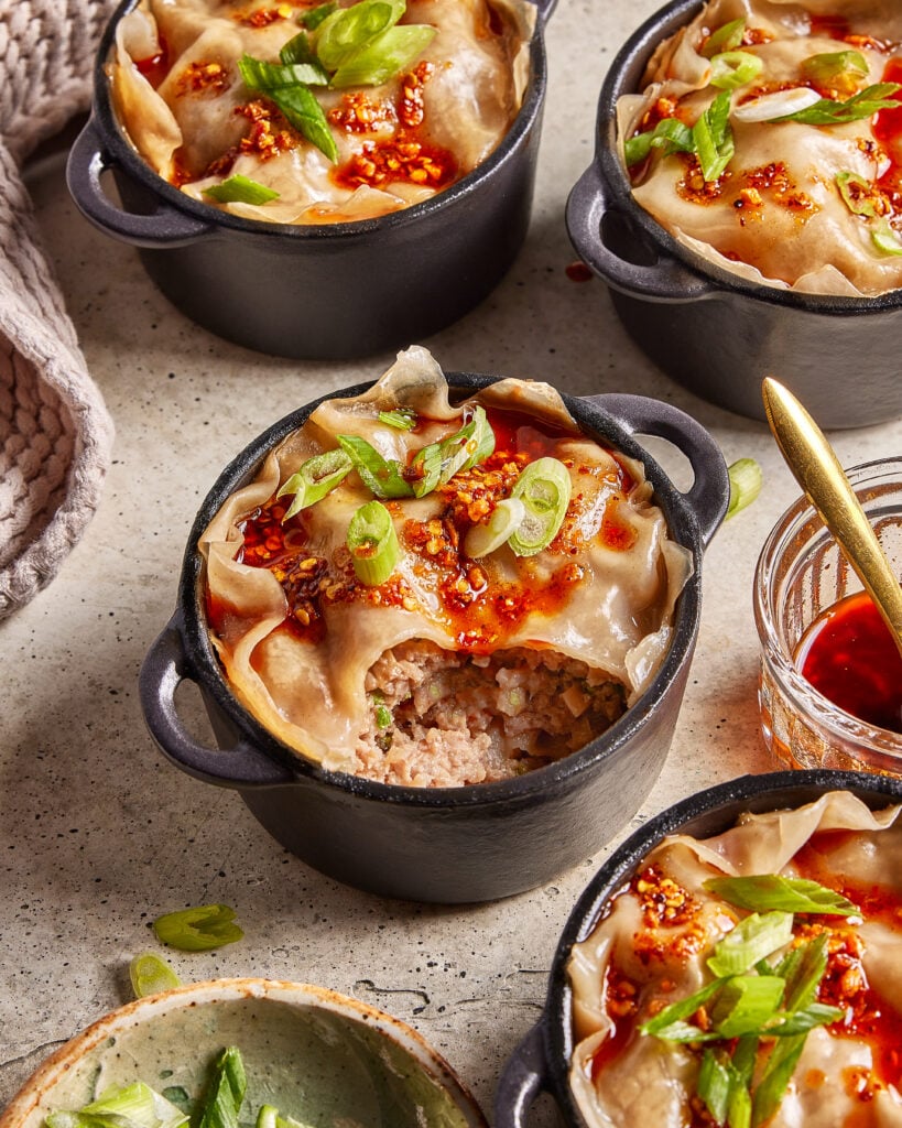 Four small black ramekins filled with dumpling pot pies are topped with chili oil, sliced green onions, and one is cut open to reveal a savory meat filling. A small bowl of sauce and spoon are nearby on a light surface.