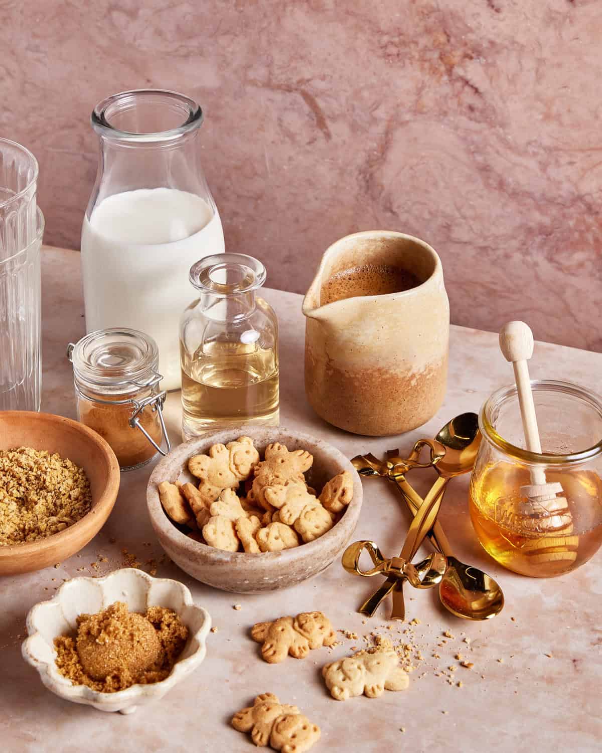 A tabletop scene with milk, honey, a small glass bottle of oil, brown sugar, crushed nuts, bear-shaped cookies, gold spoons, and a ceramic jug, all arranged on a pink marble surface.