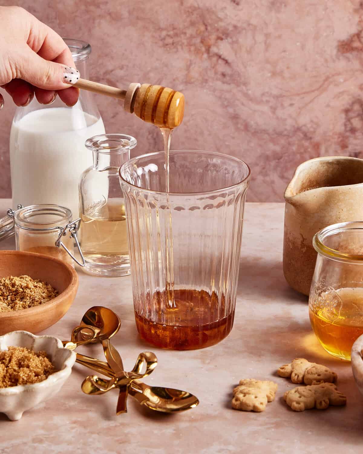 A hand drizzles honey into a clear glass surrounded by milk, brown sugar, honey, a small pitcher, golden spoons, and animal-shaped cookies on a light marble surface.