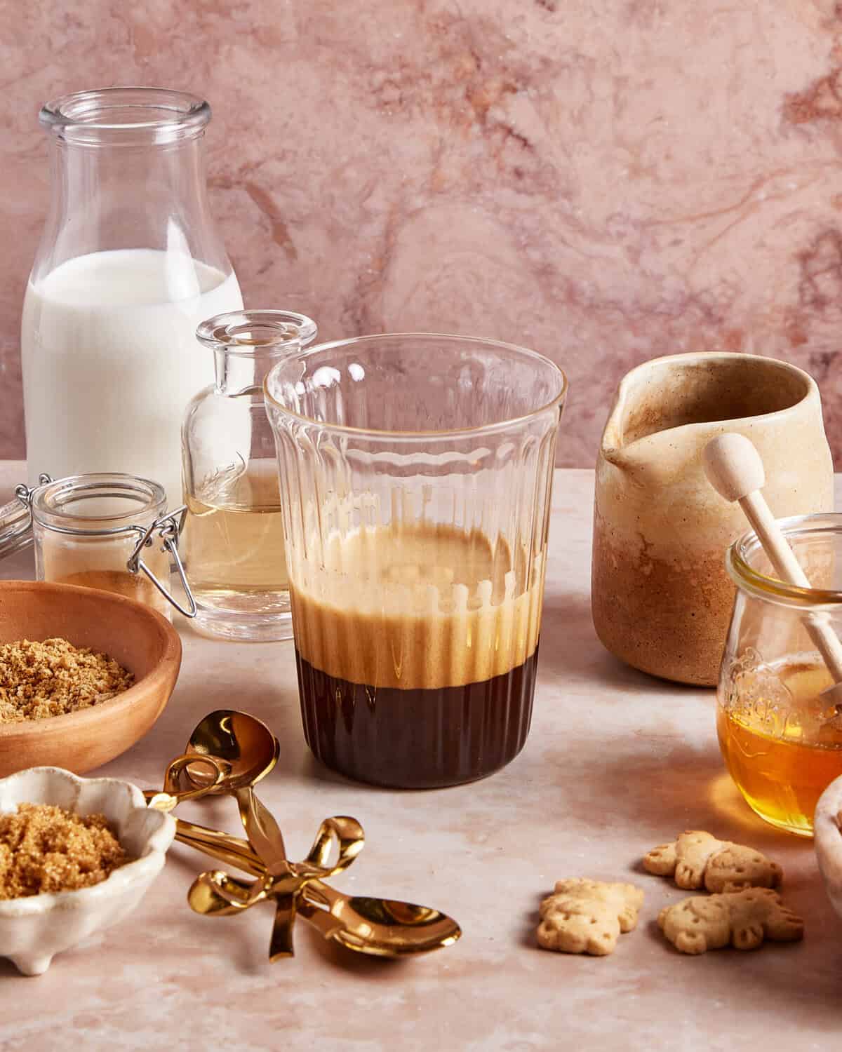 A glass of espresso with foam sits on a marble counter, surrounded by milk, honey, brown sugar, small biscuits, a gold spoon, and various jars, creating a cozy coffee setup.