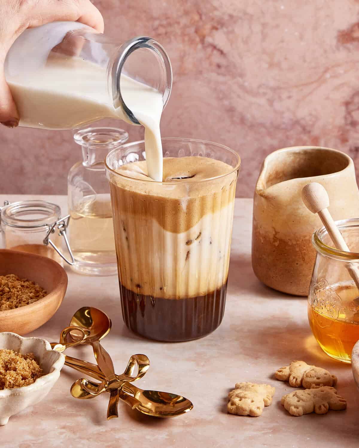 A hand pours milk into a glass of iced coffee with foam. Nearby are jars of brown sugar and honey, a honey dipper, gold spoons, and small bear-shaped cookies on a light marble surface.