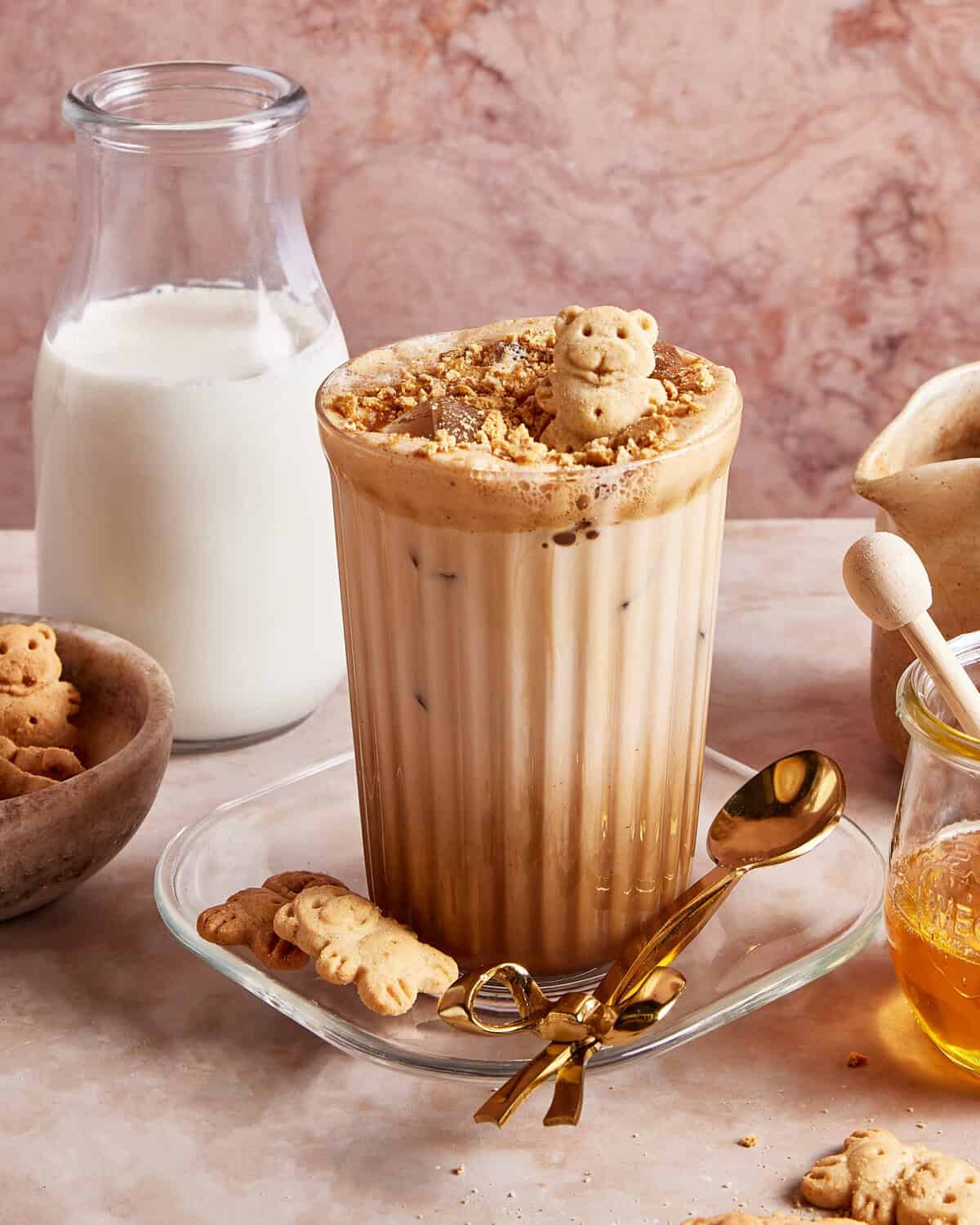 A glass of iced coffee topped with whipped foam and a bear-shaped cookie, surrounded by mini bear cookies, a jar of milk, a jar of honey with a dipper, and a golden spoon on a glass plate.
