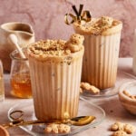 Two iced lattes in ribbed glasses topped with crushed cookies and bear-shaped biscuits, served with gold spoons. Honey, a small jug, more bear biscuits, and a bowl with cookie crumbs surround the drinks.