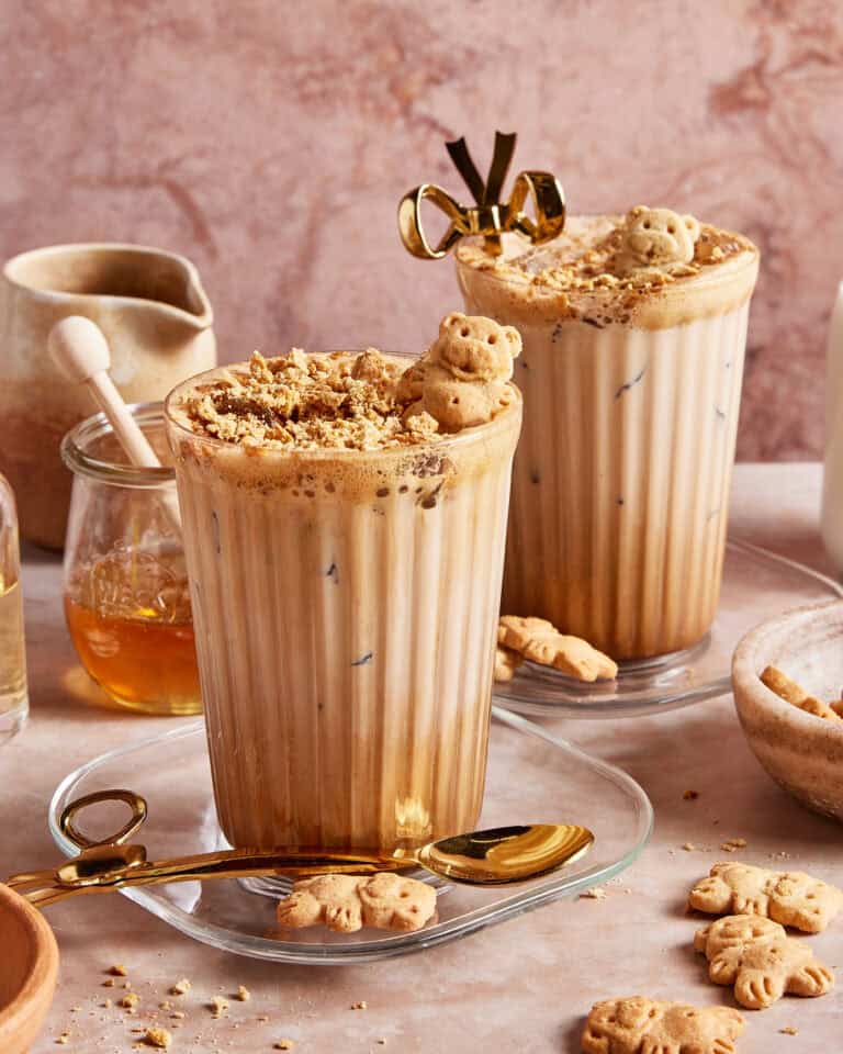 Two iced lattes in ribbed glasses topped with crushed cookies and bear-shaped biscuits, served with gold spoons. Honey, a small jug, more bear biscuits, and a bowl with cookie crumbs surround the drinks.