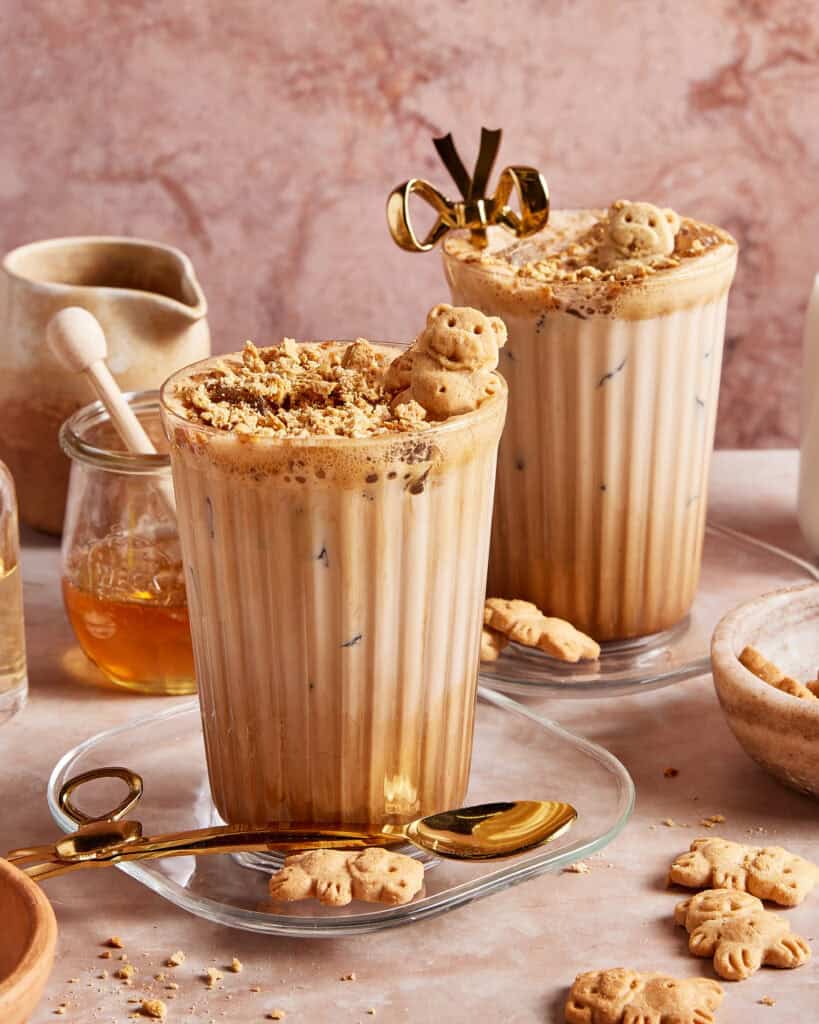 Two iced lattes in ribbed glasses topped with crushed cookies and bear-shaped biscuits, served with gold spoons. Honey, a small jug, more bear biscuits, and a bowl with cookie crumbs surround the drinks.