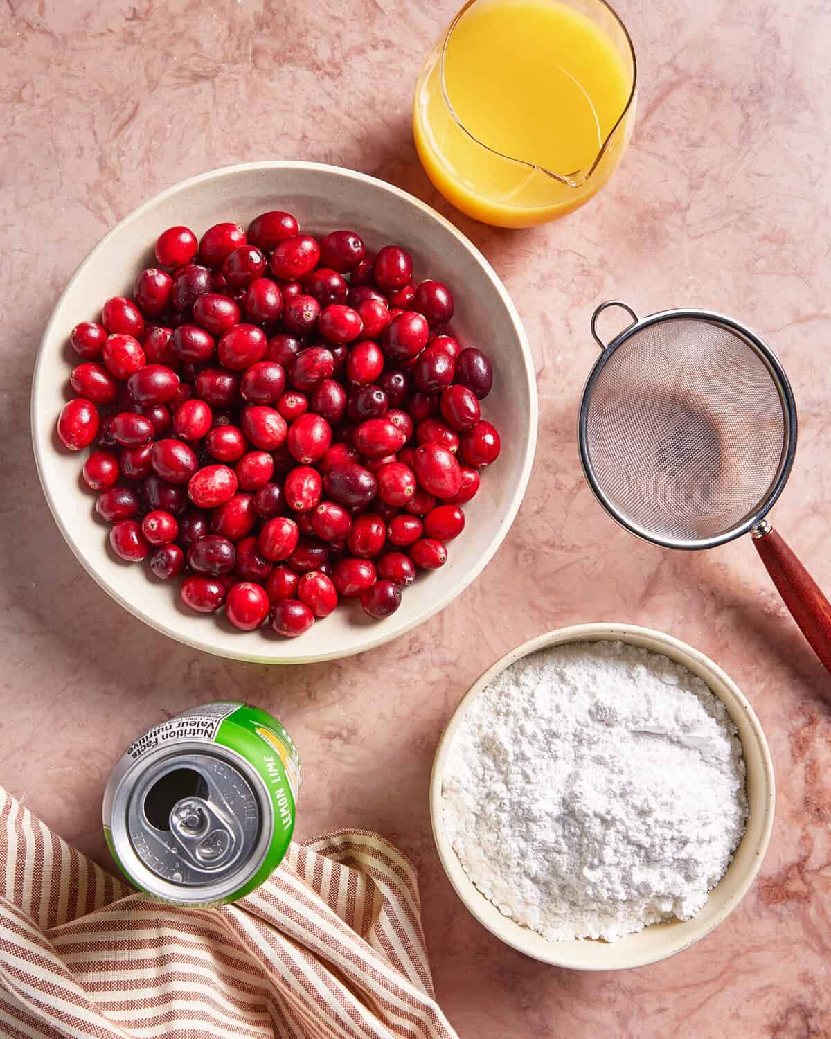 A bowl of fresh cranberries, a glass of orange juice, a mesh strainer, a bowl of powdered sugar, and an opened can of lemon-lime soda on a pink marble surface with a striped cloth nearby.