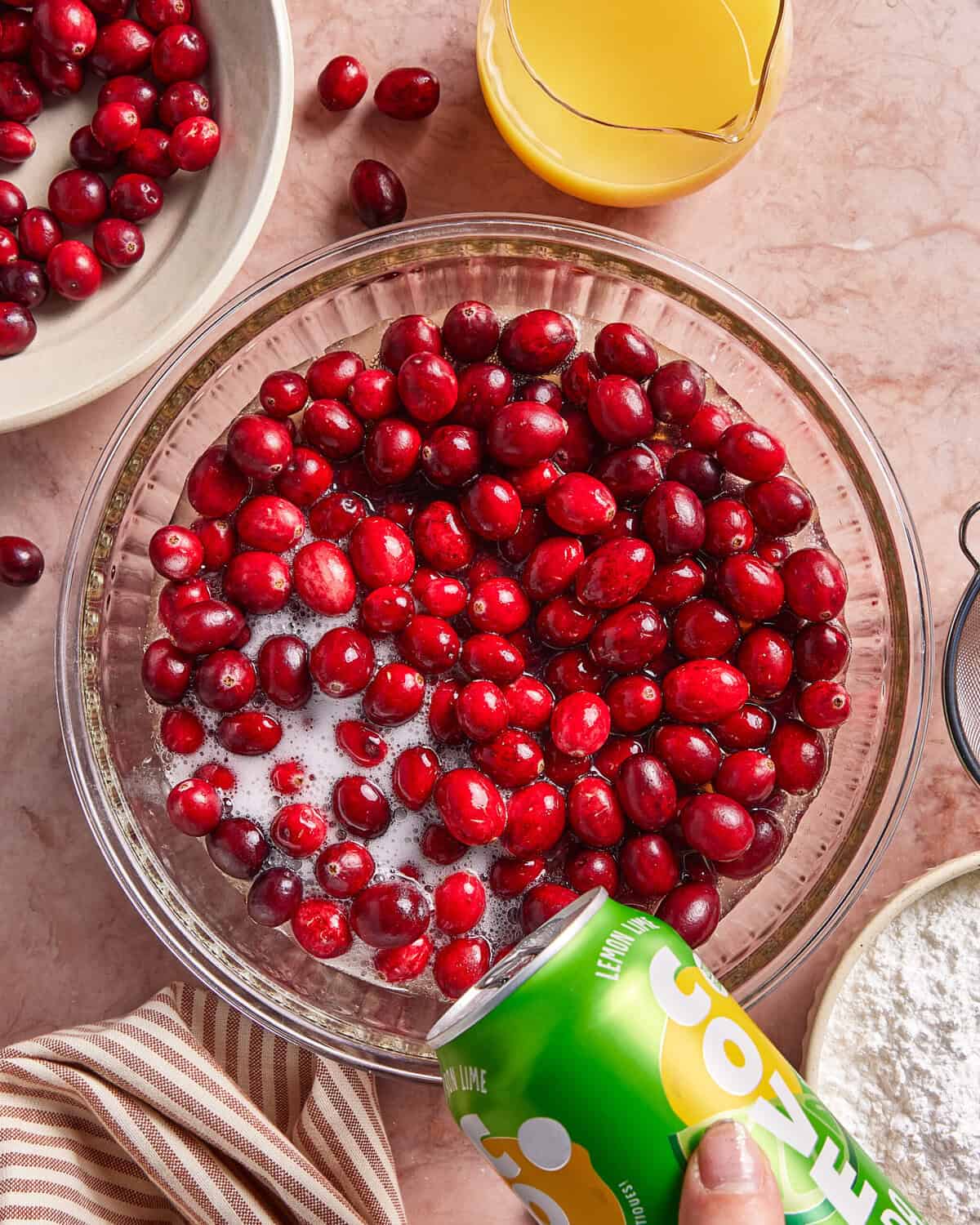 A hand pours a can of lemon-lime soda over fresh cranberries in a glass bowl. Nearby are a bowl of cranberries, orange juice in a glass, and a bowl of powder, all on a pink countertop.