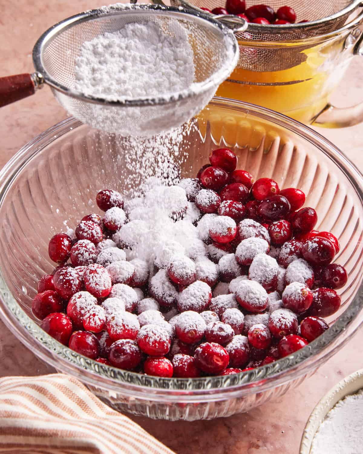 A glass bowl filled with fresh cranberries being dusted with powdered sugar through a fine mesh strainer, with a strainer and bowl of juice in the background.