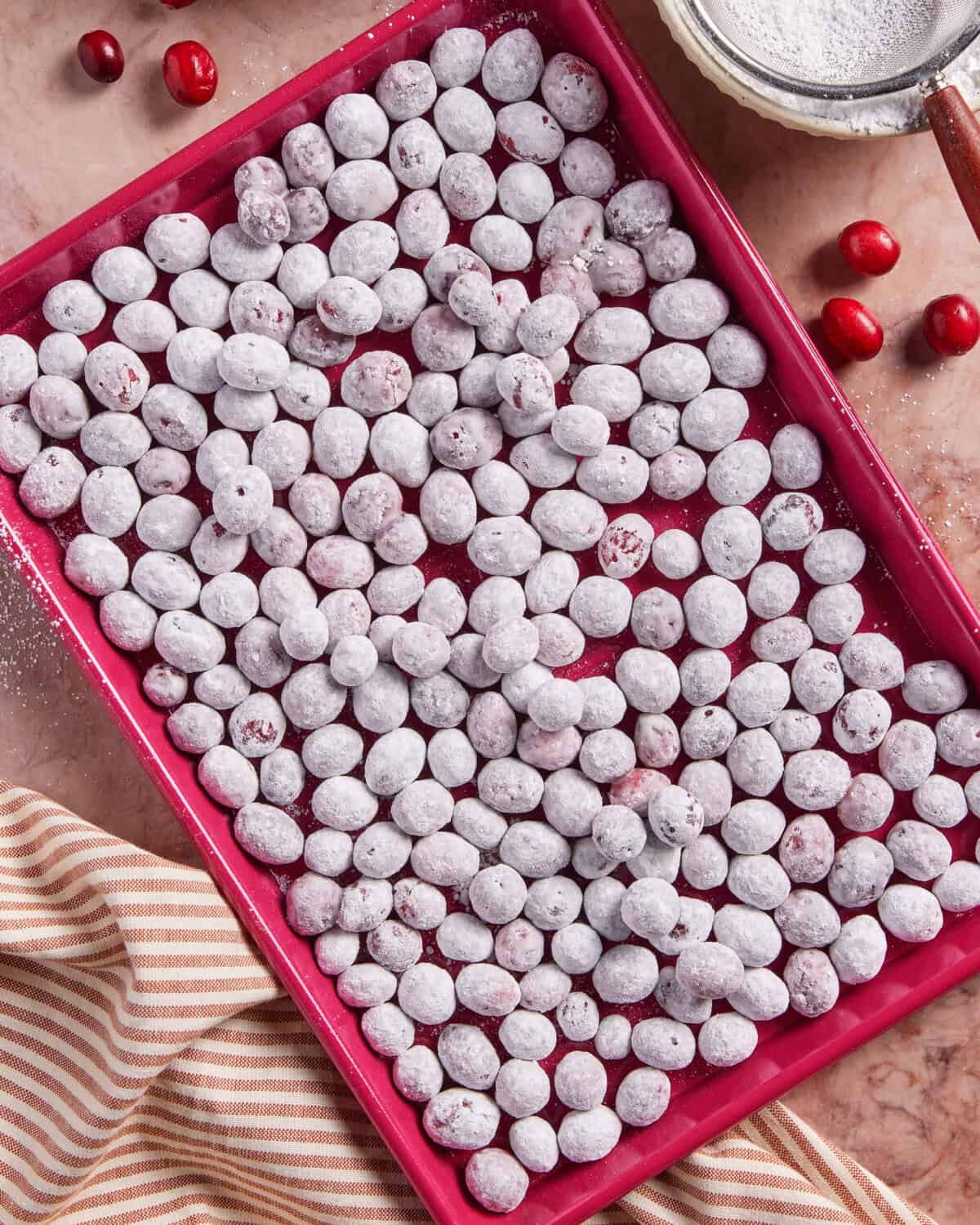 A red tray filled with sugared cranberries coated in white sugar, surrounded by loose cranberries, a mesh strainer with powdered sugar, and a striped kitchen towel on a marble surface.