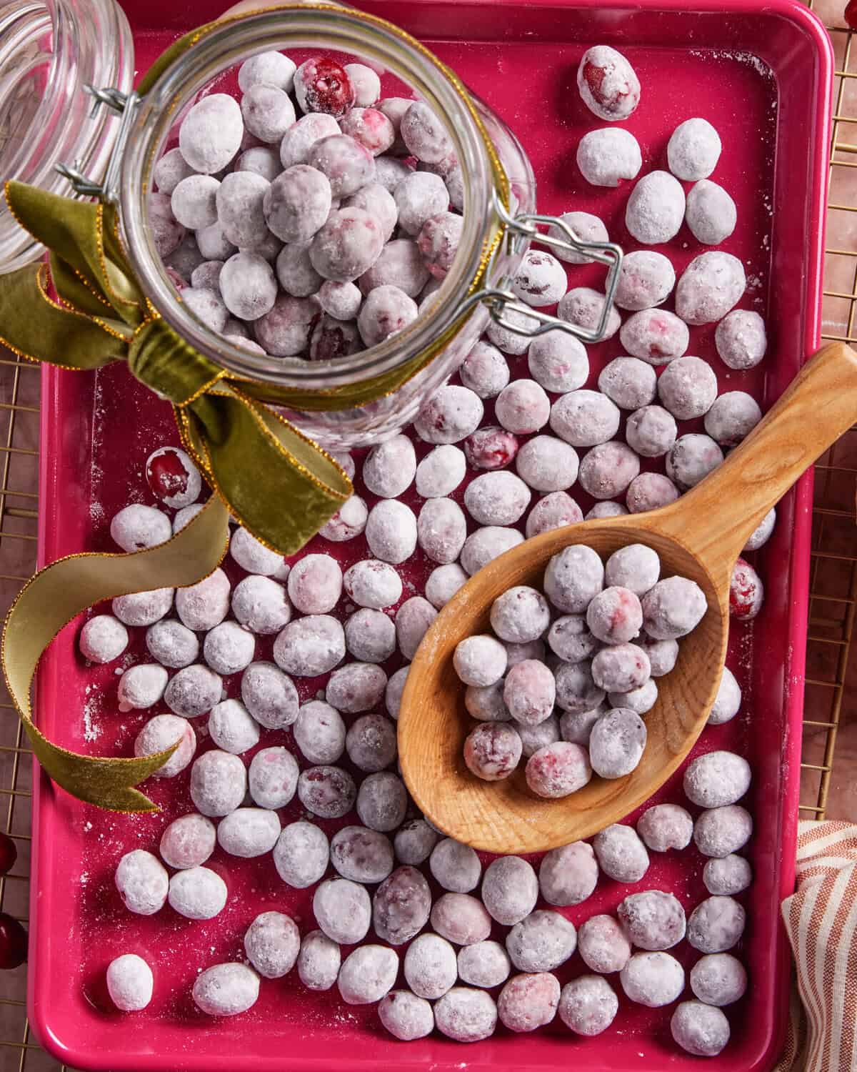 Sugared cranberries scattered on a red tray, with some in a glass jar tied with a green ribbon and others in a wooden spoon, creating a festive and sweet presentation.