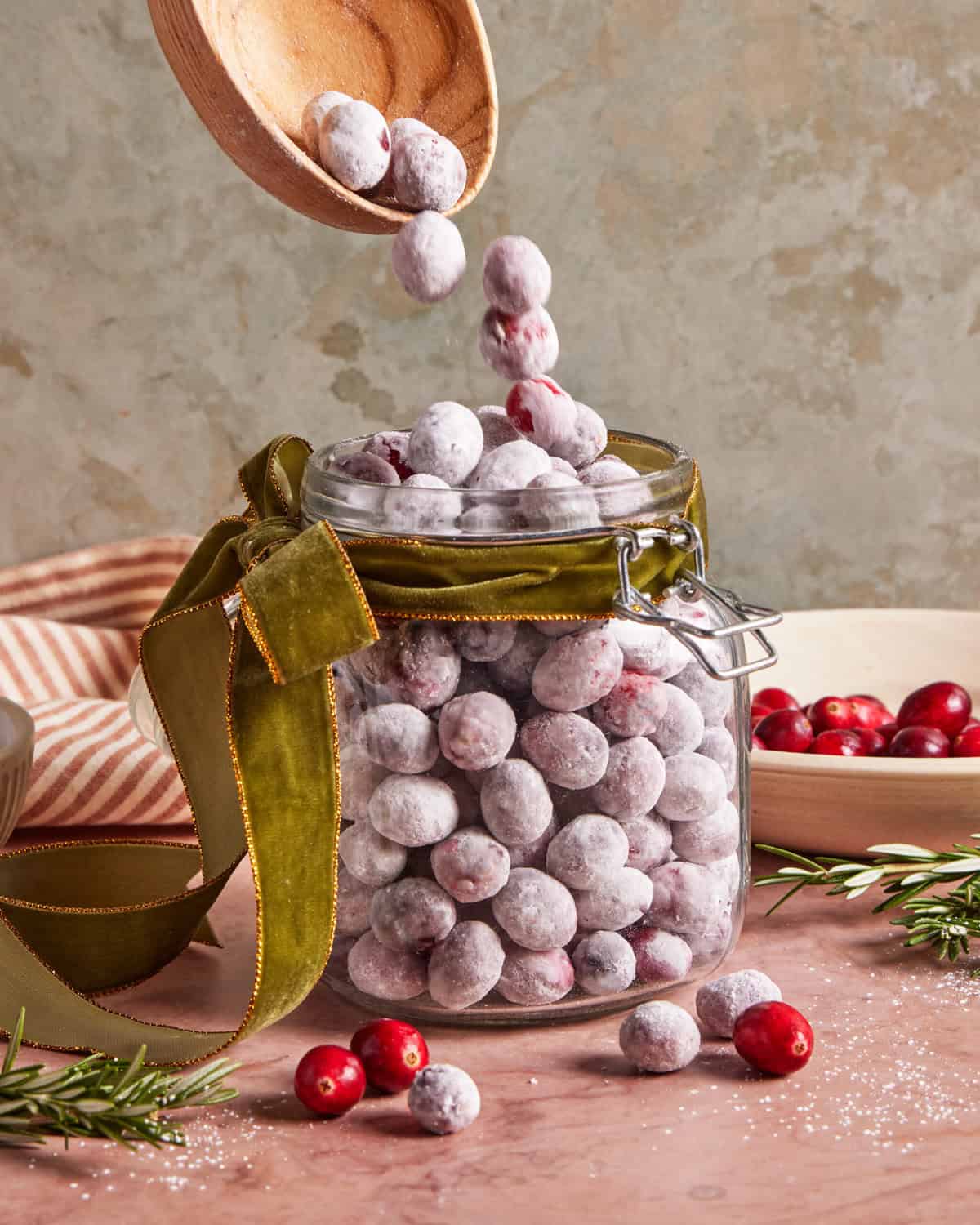A glass jar filled with powdered sugar-coated cranberries, decorated with a green velvet ribbon. A wooden spoon drops more cranberries into the jar. Loose cranberries and rosemary sprigs sit on the table nearby.