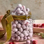 A glass jar filled with sugared cranberries, topped with more cranberries and tied with a green velvet ribbon. Loose cranberries and rosemary sprigs are scattered around the jar on a pink surface.