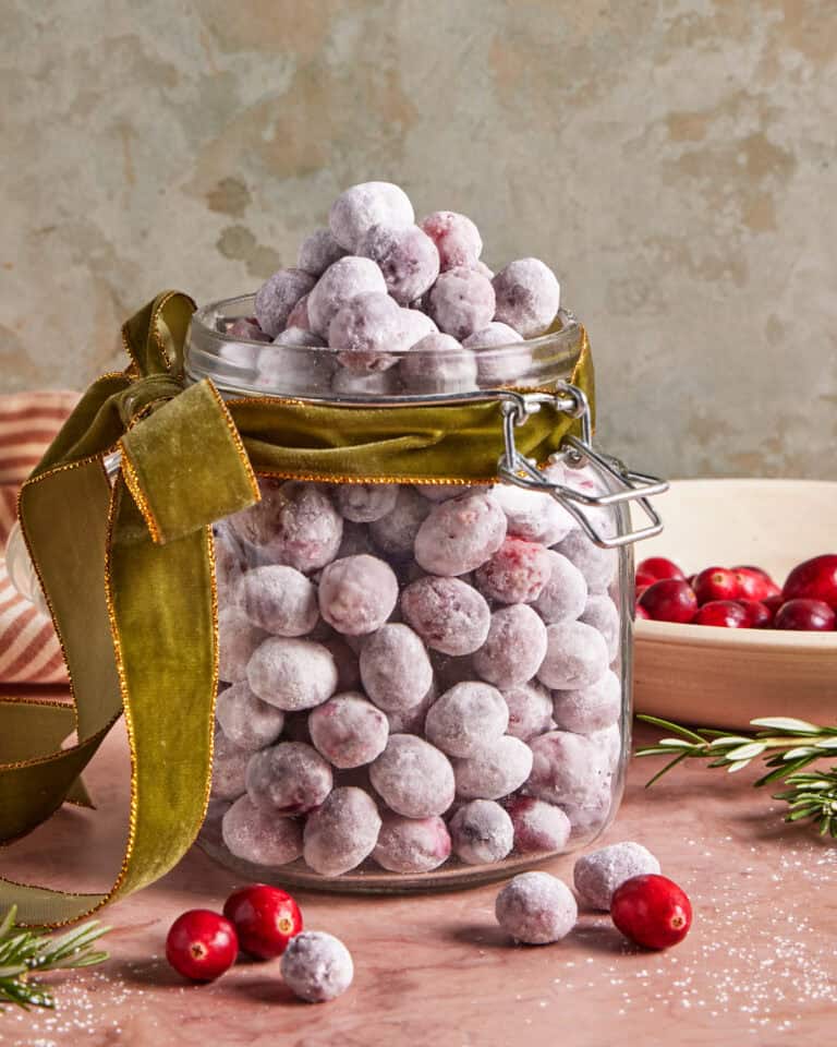 A glass jar filled with sugared cranberries, topped with more cranberries and tied with a green velvet ribbon. Loose cranberries and rosemary sprigs are scattered around the jar on a pink surface.