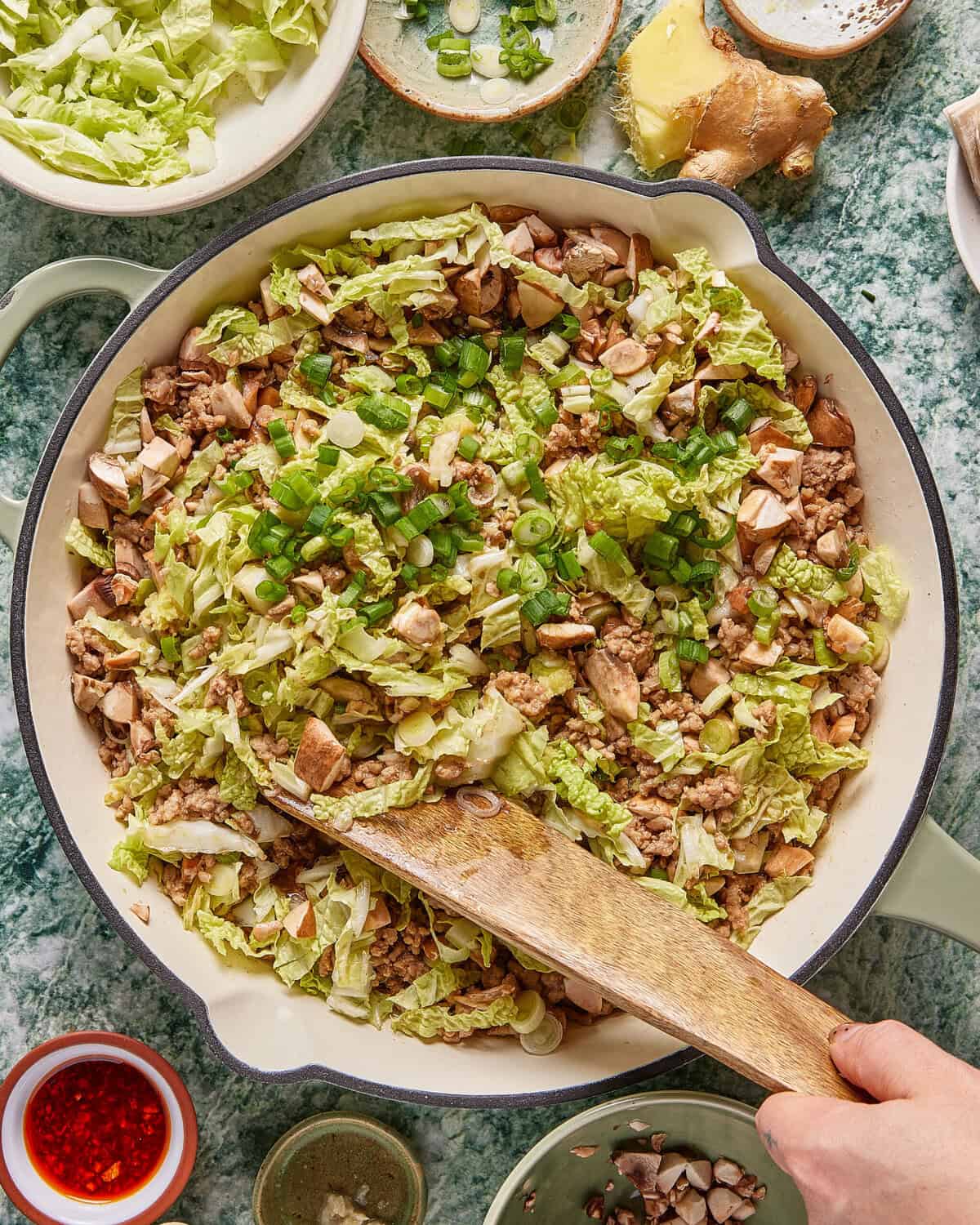 A large pan filled with chopped cabbage, ground meat, mushrooms, and green onions being stirred with a wooden spatula. Surrounding the pan are small bowls of sauces and chopped ingredients on a green countertop.