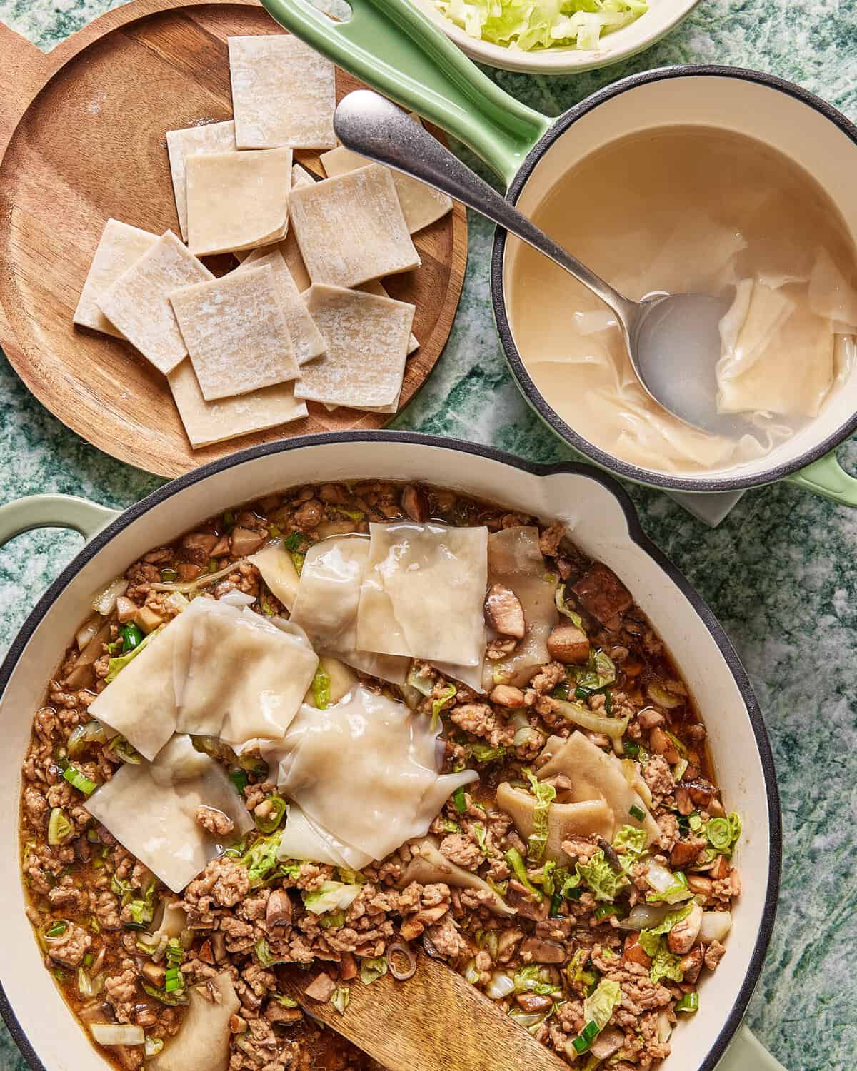 A pot of cooked ground meat and vegetables topped with square wonton wrappers, a bowl of soaked wonton wrappers with a spoon, and a wooden plate with dry wonton wrappers, all on a green marble countertop.