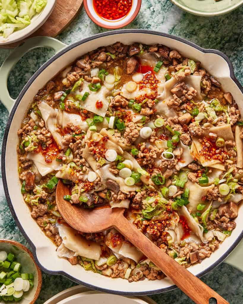 A large skillet is filled with a noodle dish containing ground meat, mushrooms, and vegetables, topped with sliced green onions and red chili oil. A wooden spoon rests in the pan, ready for serving.