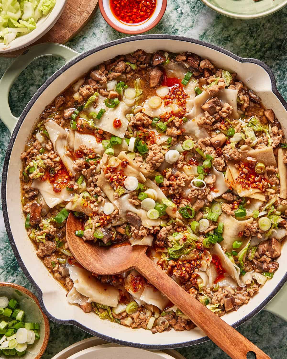 A large skillet is filled with a noodle dish containing ground meat, mushrooms, and vegetables, topped with sliced green onions and red chili oil. A wooden spoon rests in the pan, ready for serving.