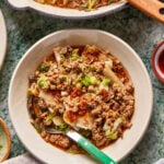 A bowl of noodle soup topped with ground meat, chopped green onions, mushrooms, and chili oil, with a green-handled spoon resting in the bowl. A pan of more soup is partially visible above.