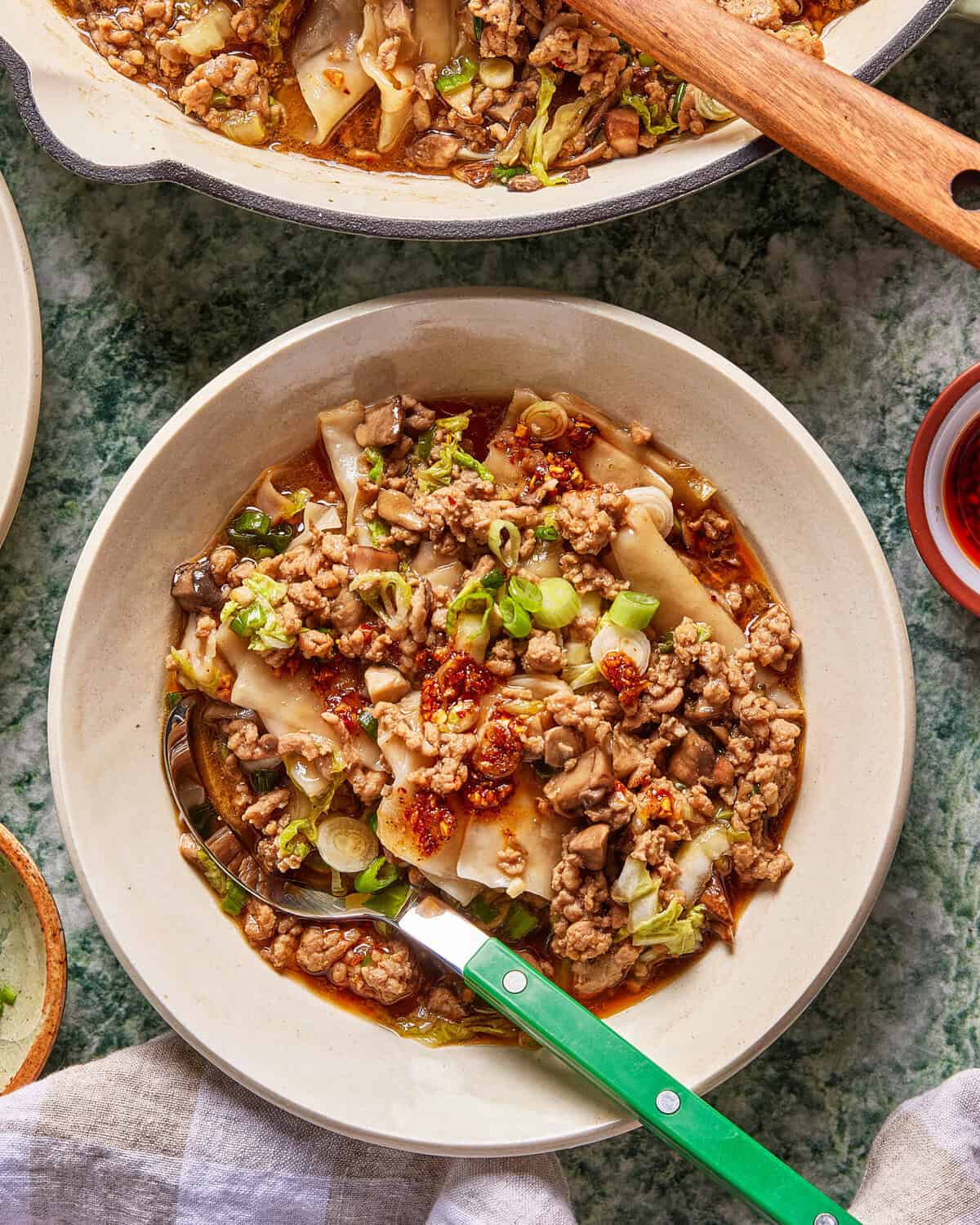 A bowl of noodle soup topped with ground meat, chopped green onions, mushrooms, and chili oil, with a green-handled spoon resting in the bowl. A pan of more soup is partially visible above.