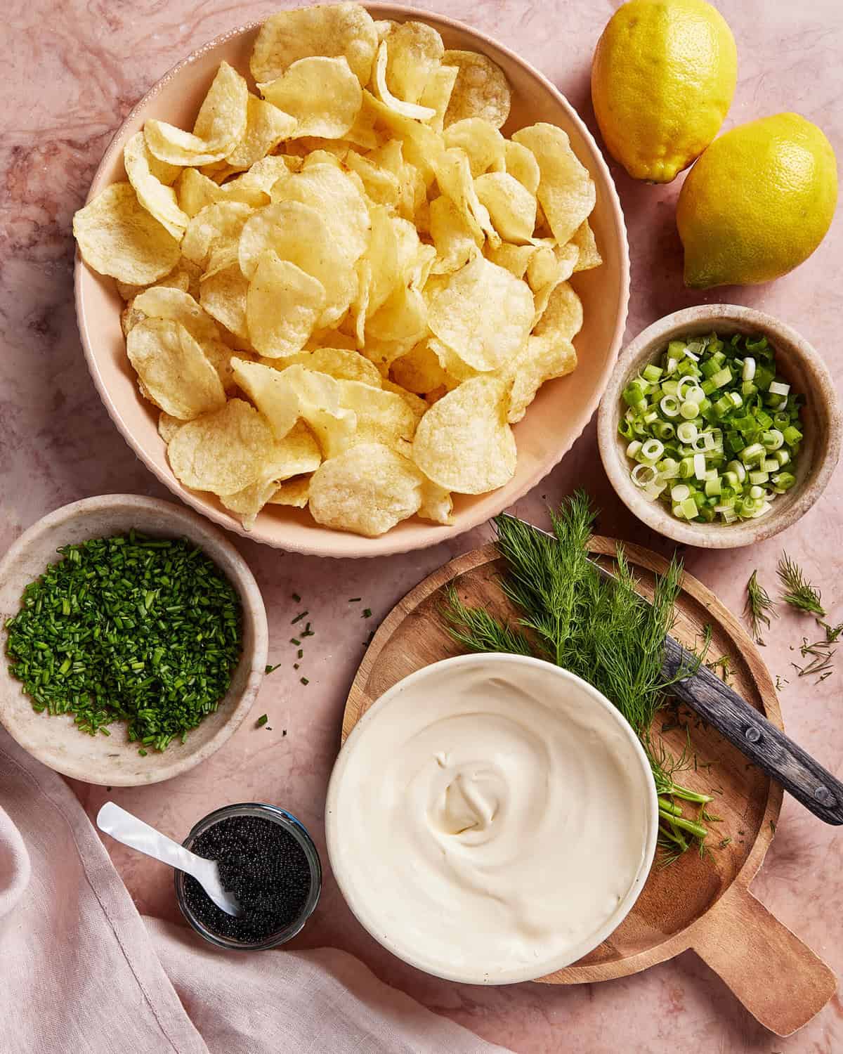 A bowl of potato chips, a bowl of mayonnaise or sour cream, chopped green onions, fresh dill, chopped chives, poppy seeds, and two whole lemons arranged on a pink marble surface.