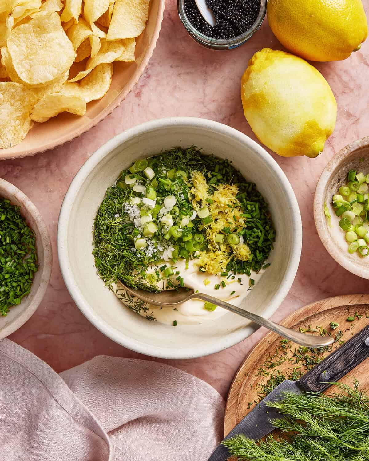 A bowl containing mayonnaise, chopped green onions, dill, chives, lemon zest, and salt with a spoon inside. Surrounding the bowl are lemons, potato chips, herbs, and a small dish of sliced green onions.