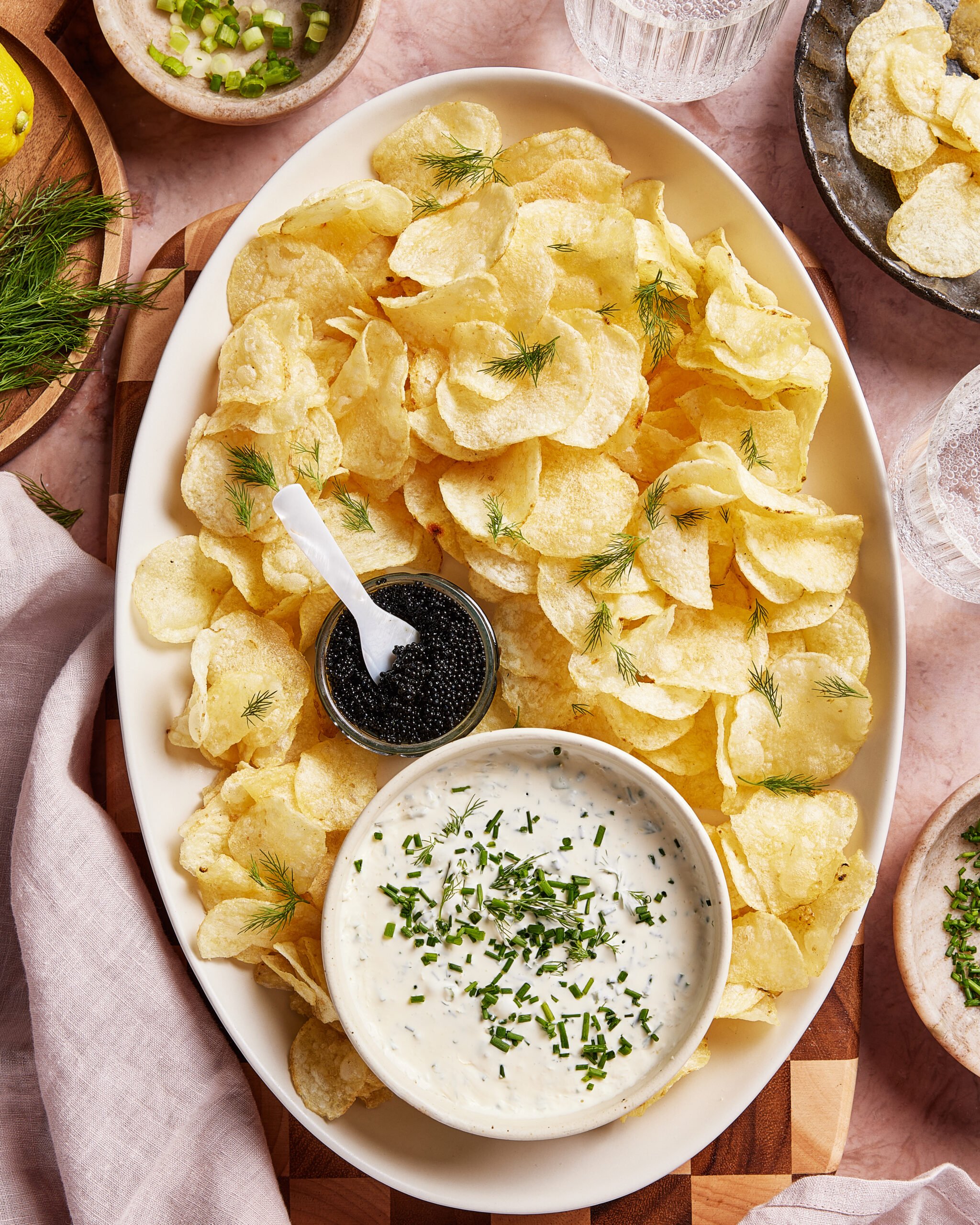 An oval platter with crispy potato chips, a small bowl of black caviar, and a larger bowl of creamy dip garnished with chopped herbs. Some chips are topped with sprigs of dill. The platter sits on a light cloth.