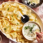 A large platter of potato chips is served with a bowl of creamy dip topped with chopped chives and a small container of black caviar. A hand dips a chip into the bowl, with fresh dill sprinkled over the chips.
