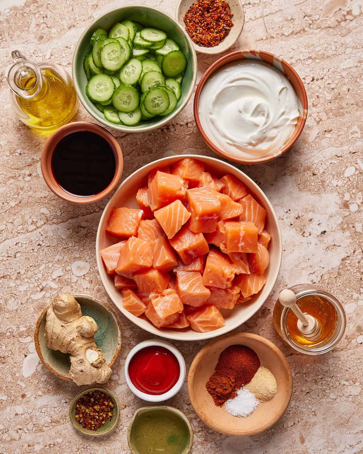 An overhead view of ingredients in bowls on a countertop, including raw salmon cubes, sliced cucumber, yogurt, olive oil, ginger, soy sauce, honey, spices, and chili flakes.