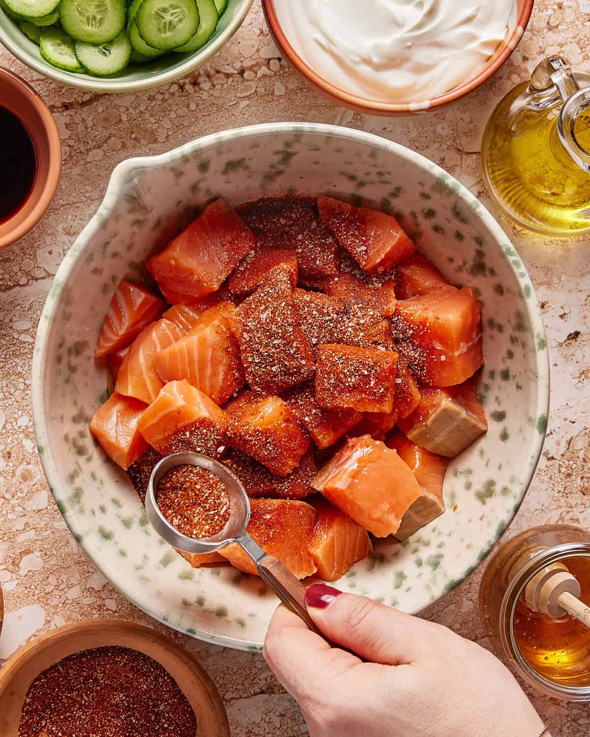 A hand sprinkles a spoonful of seasoning onto cubed raw salmon in a bowl. Surrounding the bowl are ingredients like sliced cucumbers, sour cream, olive oil, and other seasonings.