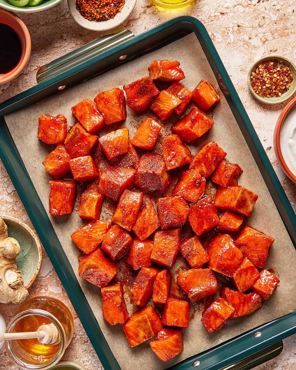 A baking tray lined with parchment paper holds roasted, seasoned sweet potato cubes. Surrounding the tray are small bowls with spices, honey, ginger, and dipping sauces on a light textured surface.