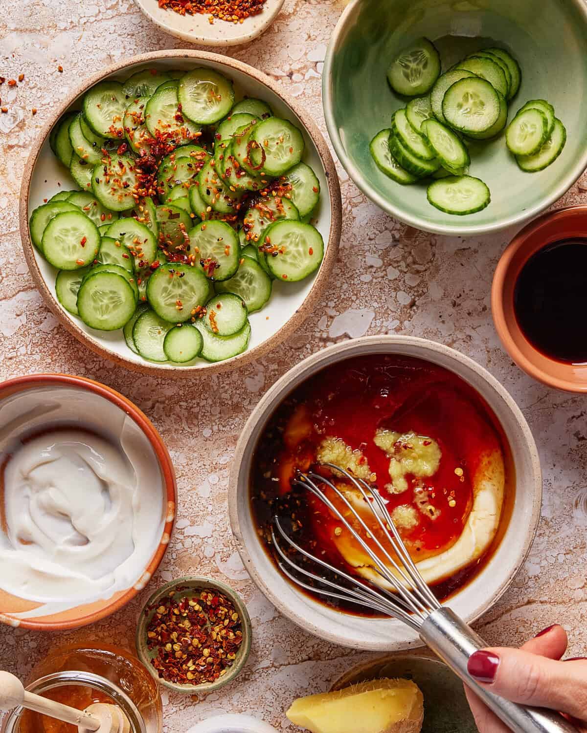 A hand whisks a red sauce in a bowl surrounded by sliced cucumbers, yogurt, soy sauce, chili flakes, and ginger on a textured surface.
