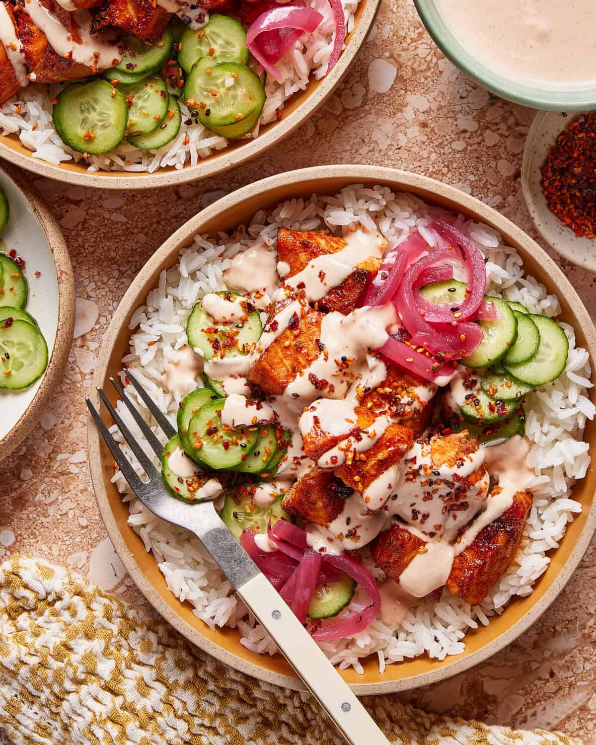 A bowl of white rice topped with grilled chicken, sliced cucumbers, pickled red onions, creamy sauce, and red pepper flakes, with a fork on the side and a woven napkin nearby.