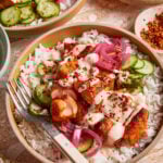 A bowl of white rice topped with seasoned chicken, sliced cucumbers, pickled onions, creamy sauce, and chili flakes, with a fork resting on the side of the bowl.