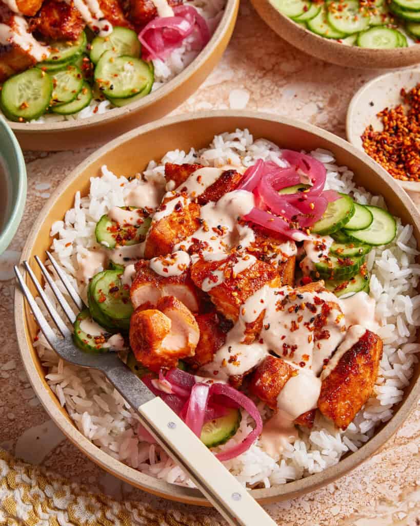 A bowl of white rice topped with seasoned chicken, sliced cucumbers, pickled onions, creamy sauce, and chili flakes, with a fork resting on the side of the bowl.