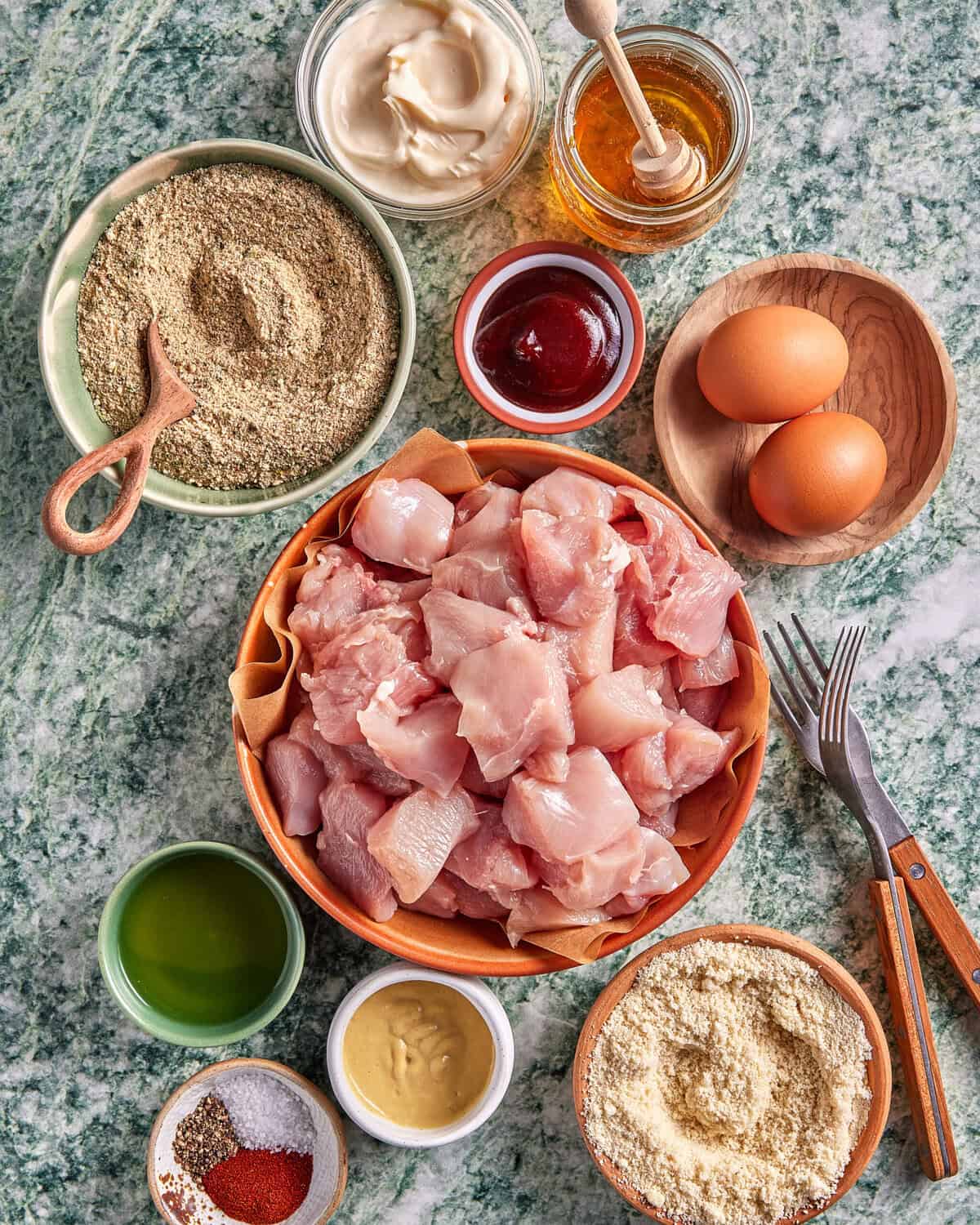 A bowl of raw, cubed chicken surrounded by eggs, breadcrumbs, flour, oil, mayonnaise, mustard, honey, ketchup, and various spices on a green marble countertop.