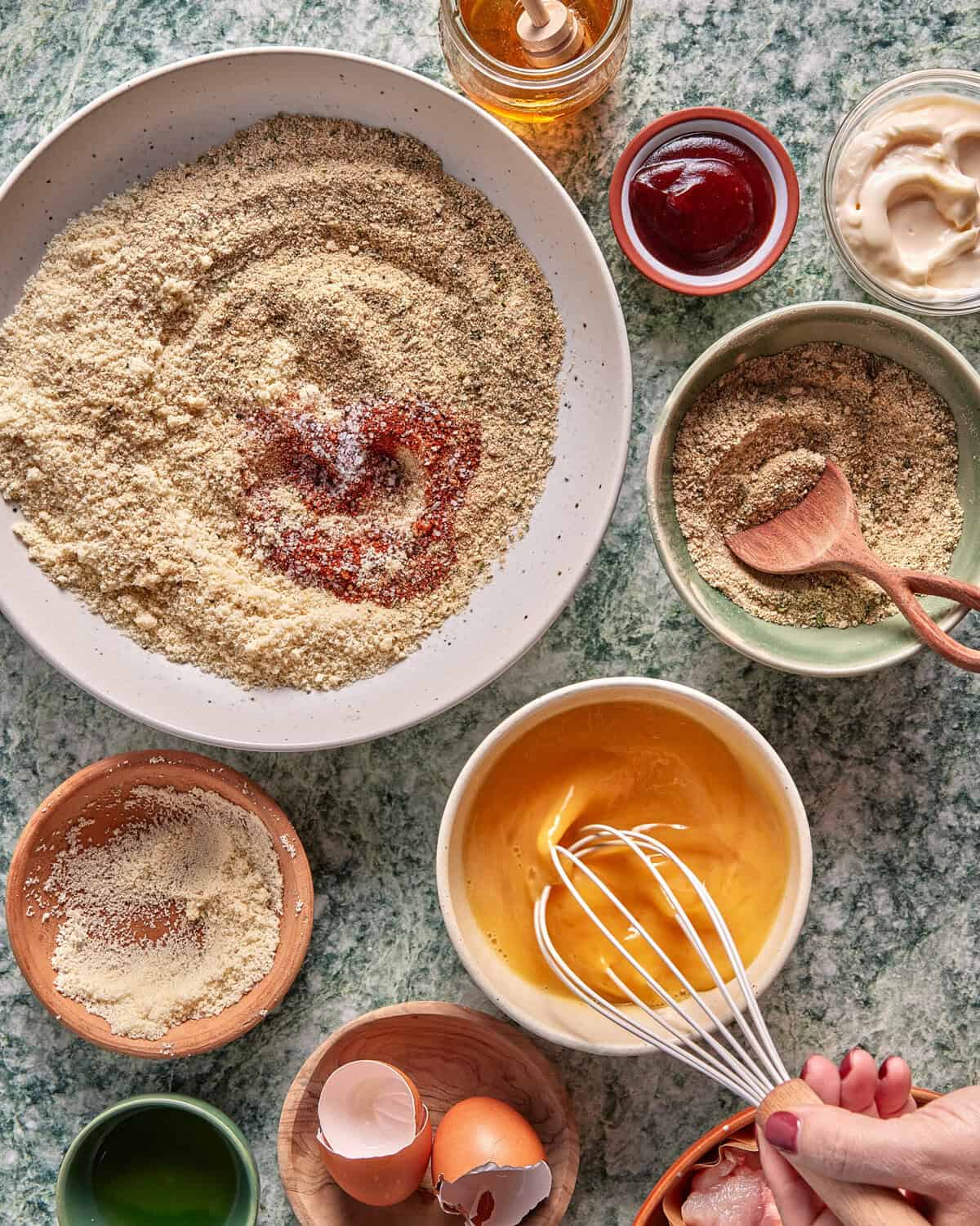 A hand whisking eggs in a bowl surrounded by bowls of breadcrumbs, sauces, eggshells, and a jar of honey on a green marble countertop, ready for breading ingredients.